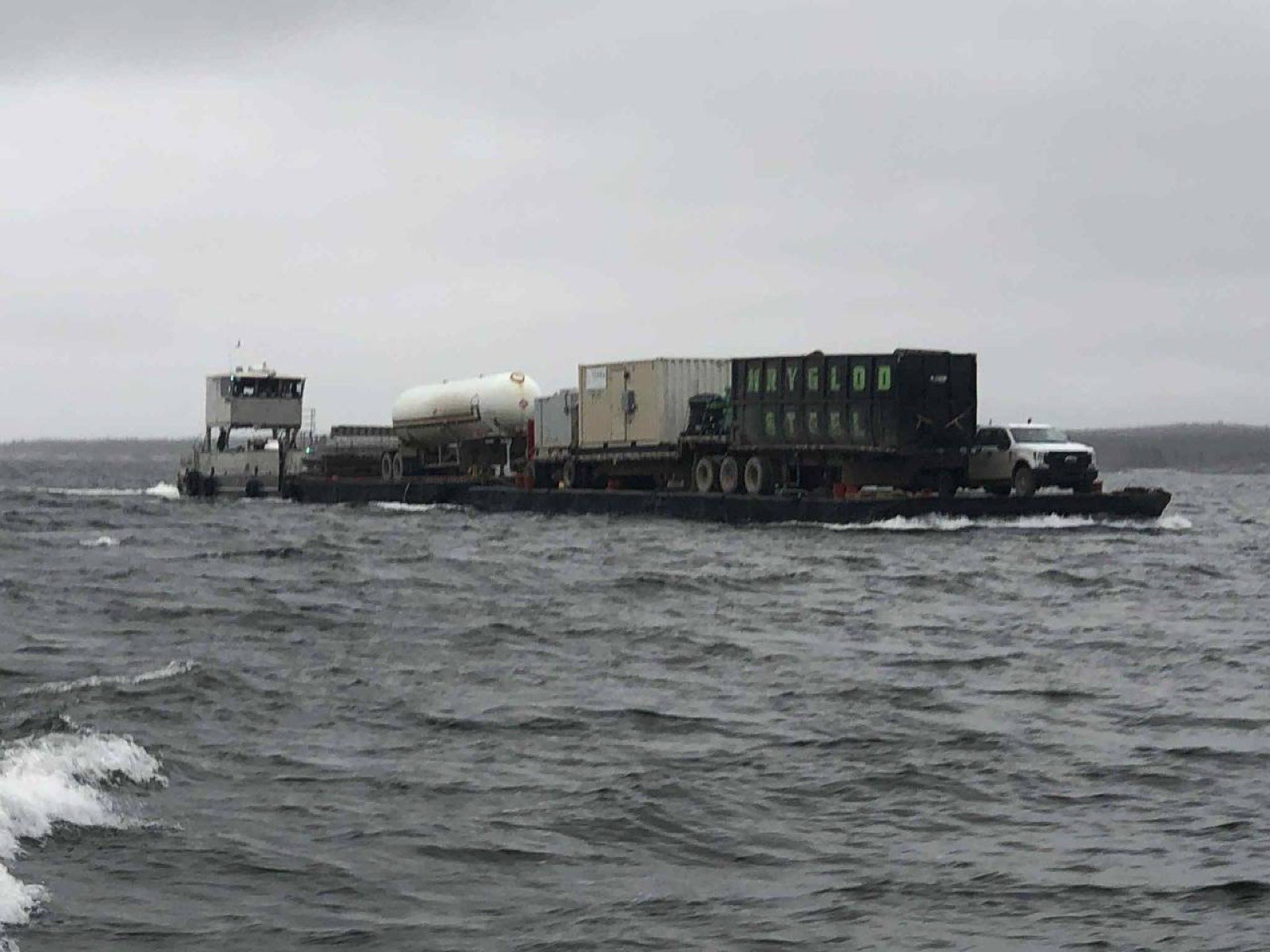 Loaded barge carrying a propane tank, shipping containers and a truck across choppy water